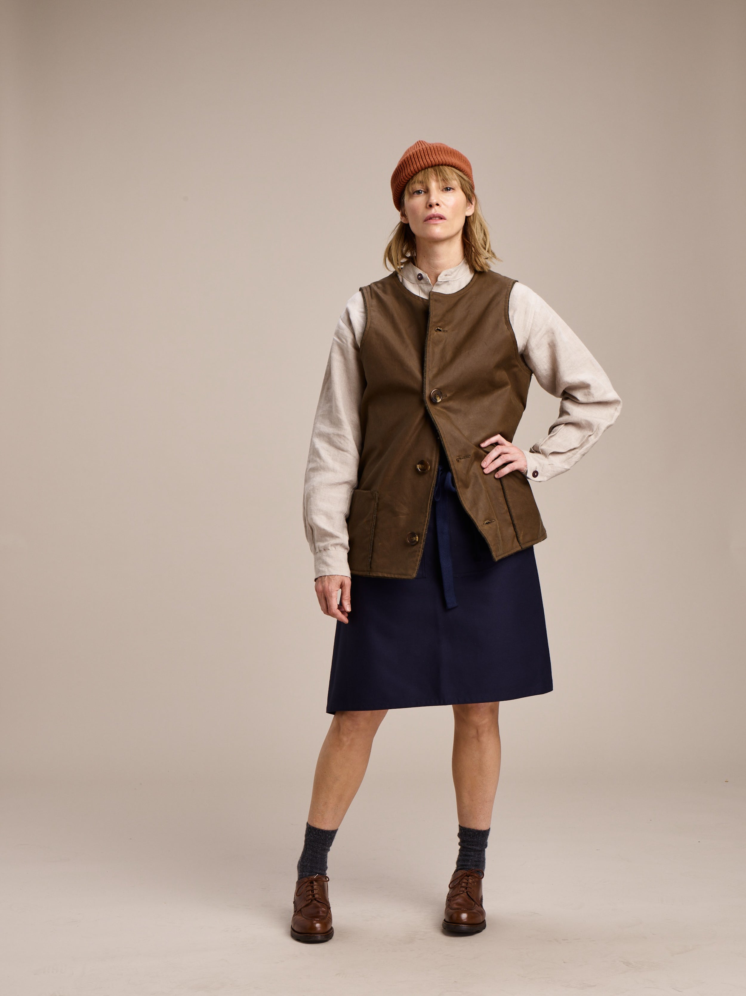 Woman wears Carrier Company Mum Skirt in Navy with norfolk Wax Cotton Jerkin in Tan and Collarless Work Shirt in Oat Linen