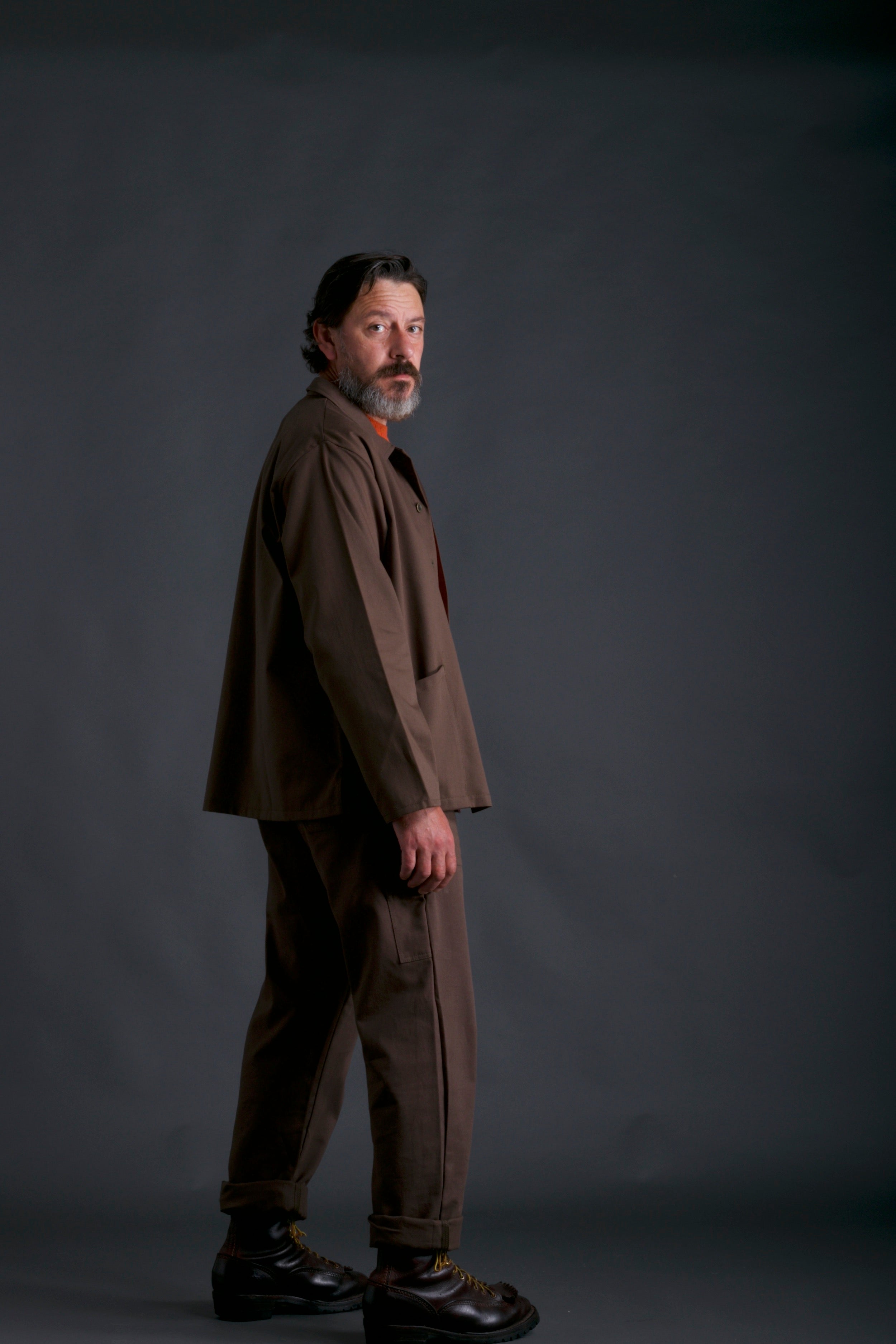 Man wears Carrier Company Men's full-length Dungarees in Olive with Shetland Lambswool Jumper in Tangerine and Norfolk Work Jacket