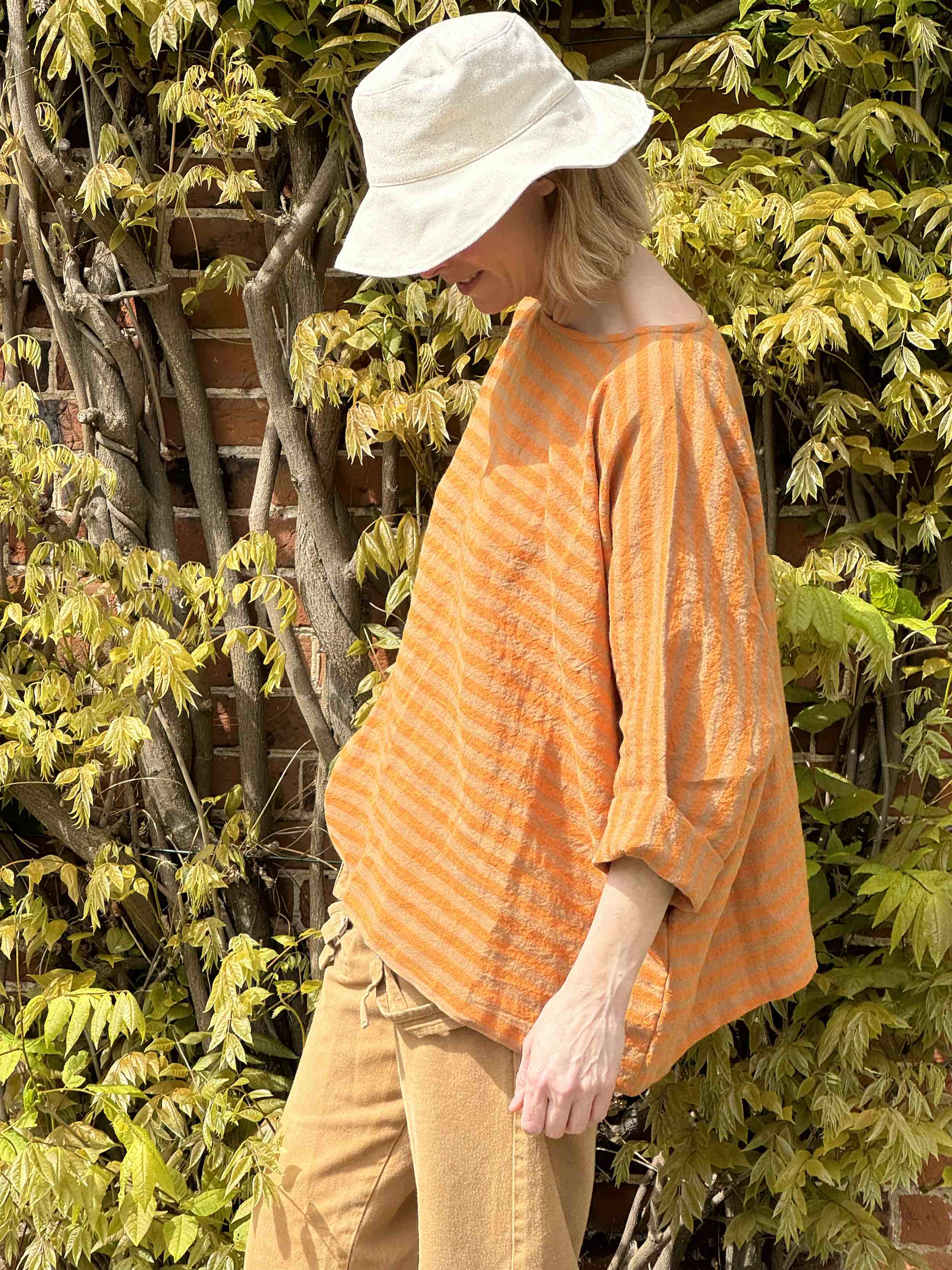 Sienna wears Orange and tan high necked boxy top with pockets and 3/4 length sleeve, with tan dungarees and sun hat